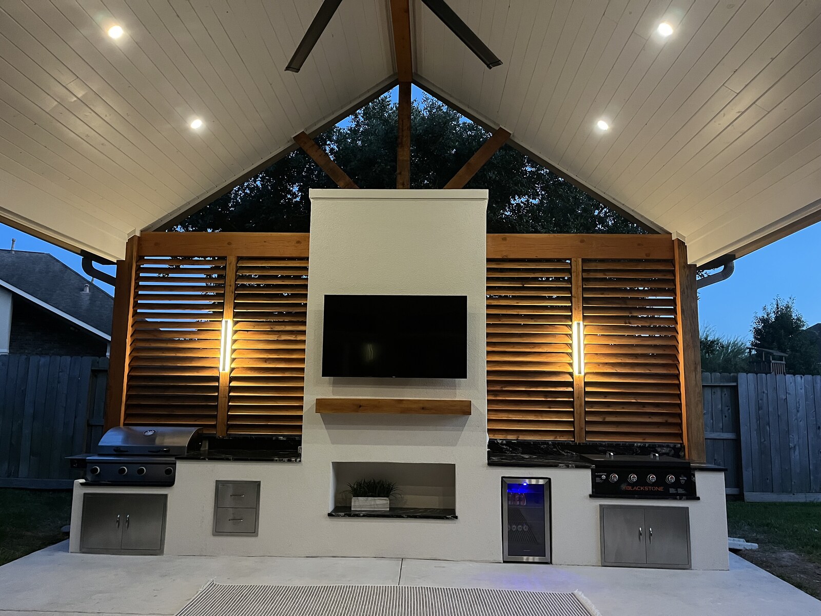 Outdoor kitchen grill islands with mounted TV and cedar shutters at dusk